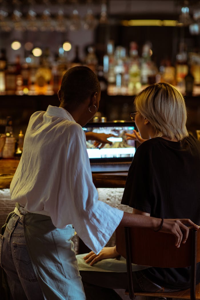 Mastering the First Impression: Your intriguing post title goes here Back view of unrecognizable black waitress in apron using netbook and pointing at screen with female colelague in casual black outfit in dark modern cozy bar