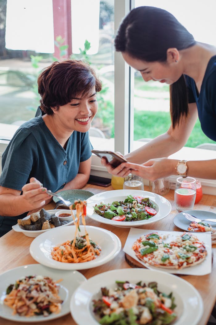 Young ethnic girlfriends sitting at table with various delicious dishes and looking at screen of mobile phone while spending time in cafe
