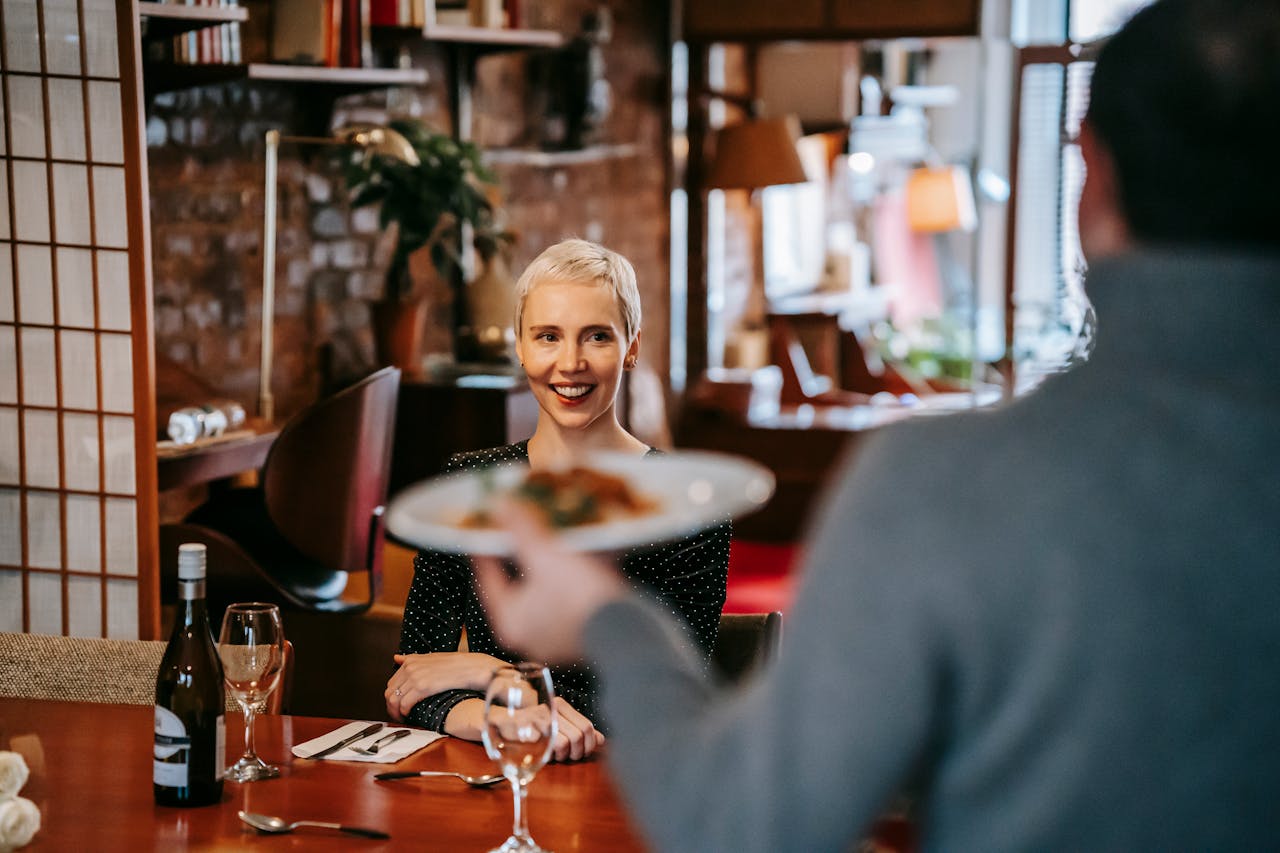 The Art of Drawing Readers In: Your attractive post title goes here Positive couple having date in bright restaurant while man serving plate with food to woman sitting in chair at table with wine bottle and glasses near cutlery with napkin